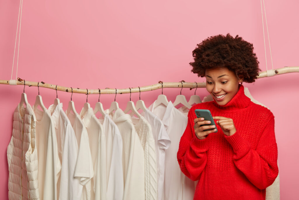 Gerente afro-americana feliz de loja online, atende clientes, digita comentários, vende nova coleção na cor branca, veste suéter vermelho de tricô, fica perto de uma arara, isolada sobre uma parede rosa. Blogueira de moda