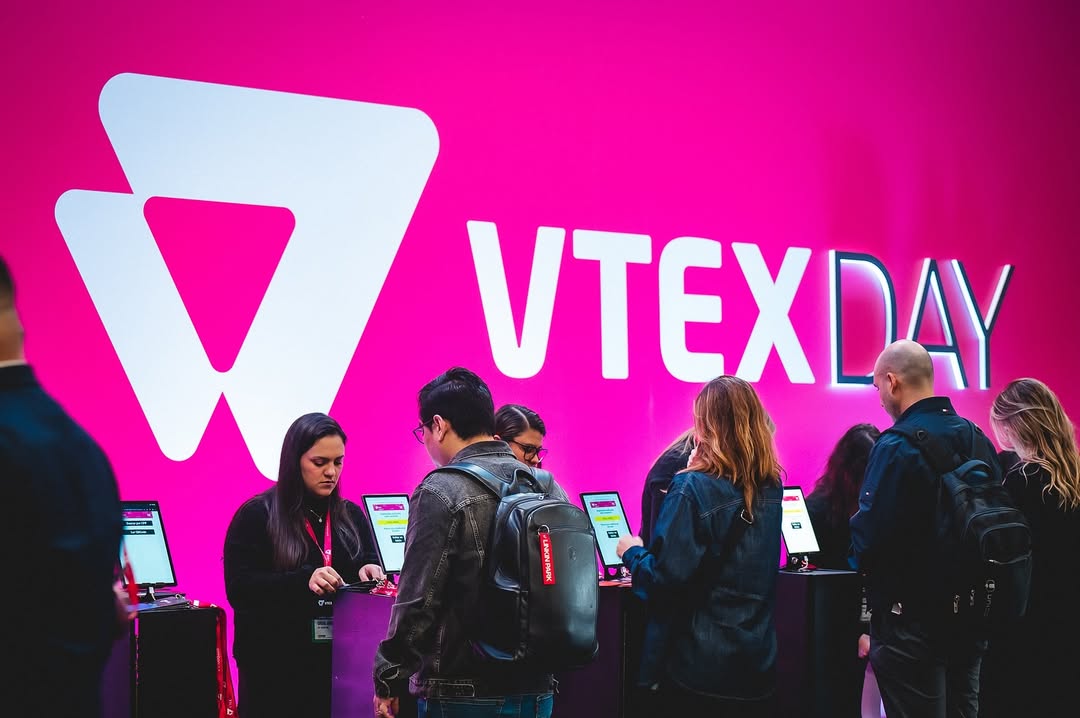 Participantes fazem check-in em totens digitais sob uma parede rosa vibrante com o logotipo e o nome "VTEX DAY" em destaque. O ambiente é moderno e movimentado, com pessoas interagindo com os dispositivos e membros da equipe auxiliando no atendimento.