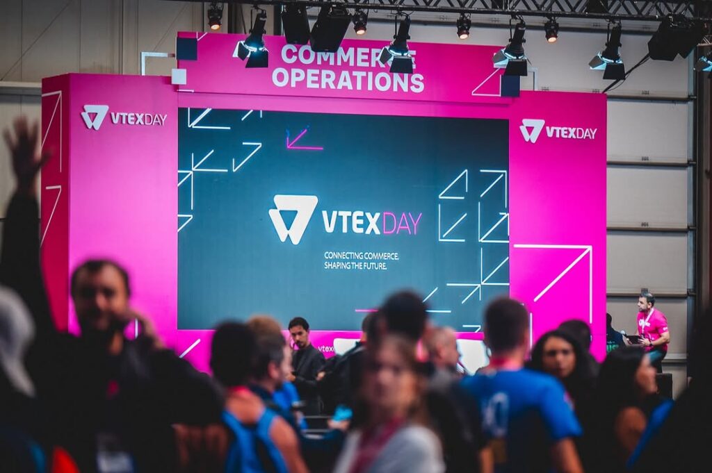 Estande do VTEX DAY com painel digital exibindo o logotipo do evento e o slogan "Connecting Commerce, Shaping the Future". O espaço é decorado em tons de rosa e preto com a inscrição "Commerce Operations" no topo, e há várias pessoas circulando e interagindo em frente ao palco, em um ambiente de conferência movimentado.