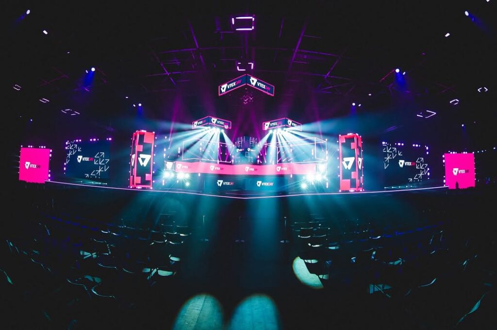 Palco iluminado com luzes de LED em tons de rosa, azul e roxo durante um evento de eSports, com estruturas tecnológicas, telões exibindo o logo da "VTV Esports", arquibancadas vazias e ambiente escuro com atmosfera futurista.