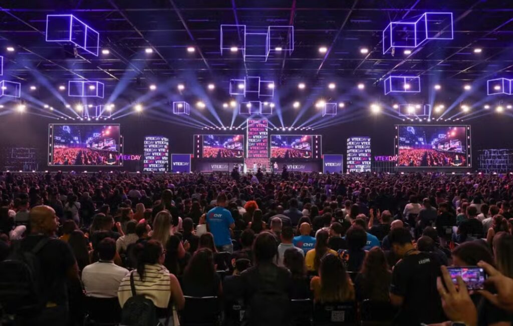 Grande público assiste a uma palestra principal no VTEX DAY, com três telões exibindo o palco ao fundo, luzes cênicas vibrantes no teto e uma estrutura moderna com iluminação em tons de roxo. O evento está lotado, com participantes atentos e registrando o momento com celulares.